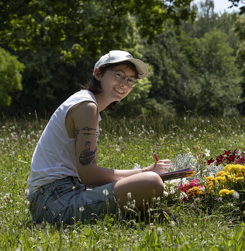 young woman sitting in the grass, flowers in front of her, paper and pencil in hand