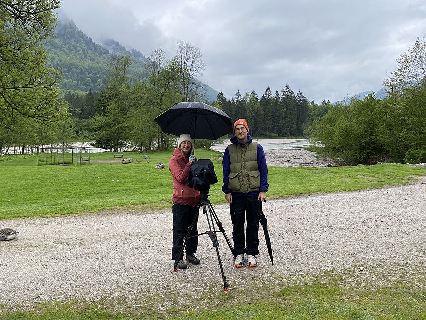 2 Personen mit professioneller Kamera mit Regenschirm auf einer Schotterstrasse im Grünen
