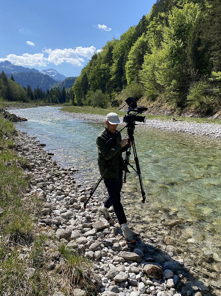 Mann trägt Kamera mit Stativ am Rande eines Flusses