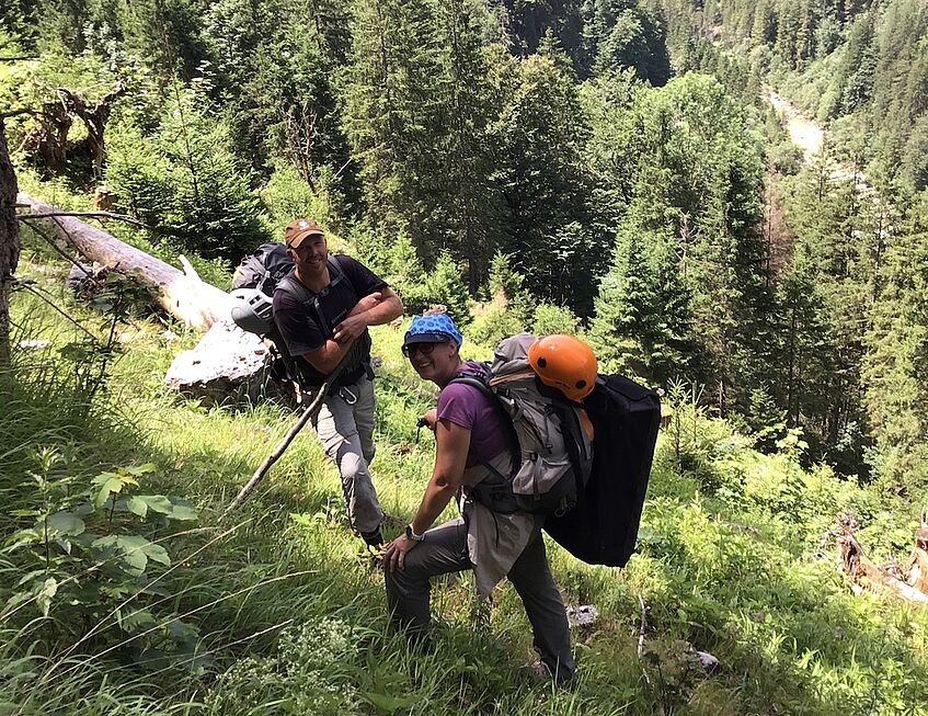 2 people with large backpacks and helmets on a hiking trail